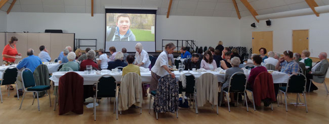 Image of people eating lunch in the Bradbury Hall with a video showing on the large screen.